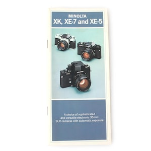 May include: A brochure featuring three Minolta cameras: XK, XE-7, and XE-5. The brochure has a blue and white color scheme with the camera models displayed on a light green background. The text on the brochure reads: "A choice of sophisticated and versatile electronic 35mm SLR cameras with automatic exposure."