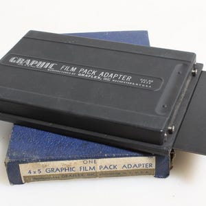 Könnte beinhalten: Ein schwarzer Graphic Film Pack Adapter, Modell 1234, hergestellt von Graflex, Inc. Der Adapter liegt auf einer blauen Box mit der Aufschrift "One 4x5 Graphic Film Pack Adapter."