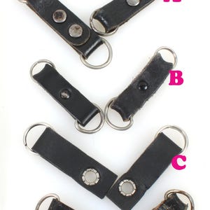 以下が含まれることがあります： 銀色の金属製のリングとリベットが付いた黒い革製のキーホルダーが4つあります。キーホルダーは縦に並べてあり、一番上のキーホルダーには「A」、2番目のキーホルダーには「B」、3番目のキーホルダーには「C」、一番下のキーホルダーには「D」とラベル付けされています。