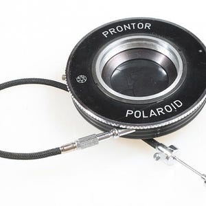 May include: A black and silver Prontor Polaroid camera lens with a black cable and a silver trigger. The lens has the word "PRONTOR" and "POLAROID" printed on it. The camera lens is on a white background.