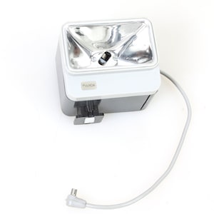 Puede incluir: Una unidad de flash blanca y negra con un reflector plateado. La unidad de flash tiene un cable con un enchufe en el extremo. La unidad de flash dice "FUJICA" en la parte delantera.