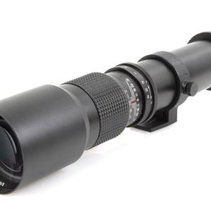 Op de afbeelding: Een zwarte telelens voor camera's met een geribbelde scherpstelring en meetmarkeringen. De lens heeft een lange, cilindrische vorm met een zonnekap aan één uiteinde. De lens is ontworpen voor professionele fotografie.