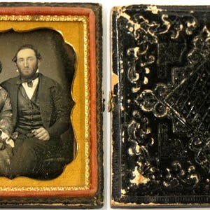 May include: An antique daguerreotype photograph in an open, hinged case. The left side displays a black and white portrait of two individuals seated together, framed by a gold-toned mat. The right side shows the black, embossed exterior case.