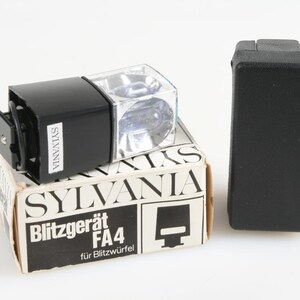 以下が含まれることがあります： 黒いSylvania Blitzgerät FA4フラッシュユニット。透明な正方形のフラッシュヘッドがあり、オリジナルの箱の上に置かれています。箱は白とタンで、黒い文字で「SYLVANIA」と「Blitzgerät FA4」と書かれています。右側には黒いケースがあります。