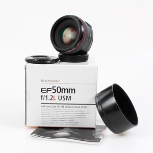 Könnte beinhalten: Ein Canon EF 50mm f/1.2L USM Objektiv mit Sonnenblende und Etui. Das Objektiv ist schwarz mit roten Akzenten und steht auf einer weißen Box. Auf der Box steht "EF 50mm f/1.2L USM".