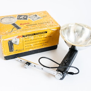 Puede incluir: Un flash Kodak Standard vintage en su caja original. La unidad de flash tiene un reflector plateado y un mango negro. La caja es amarilla con texto e imágenes negras.