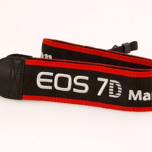 May include: A black and red camera neck strap featuring the white embroidered text EOS 7D Mark. The strap is constructed from woven fabric with black leather-like end tabs and includes adjustable black plastic buckles for secure camera attachment.