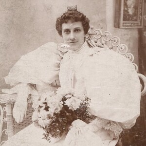 Op de afbeelding: Een sepia-getinte vintage foto van een persoon in een witte jurk met een hoge kraag, grote pofmouwen en kanten details. De persoon houdt een groot boeket bloemen vast terwijl deze op een decoratieve witte rieten stoel zit.