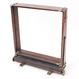 Puede incluir: Un marco de madera marrón oscuro con herrajes de latón y pequeñas ruedas. El marco es rectangular y parece plegable. La madera tiene un aspecto rico y envejecido, lo que sugiere que es vintage o antiguo.