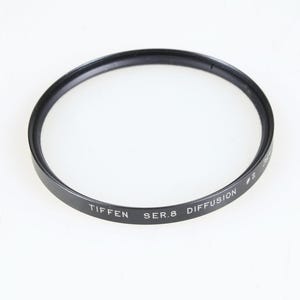 Puede incluir: Un filtro de lente de cámara circular negro Tiffen Ser.8 Diffusion #2. El marco de metal presenta texto impreso en blanco que dice Tiffen Ser.8 Diffusion #2, indicando el modelo específico y el tipo de accesorio óptico para equipo fotográfico.