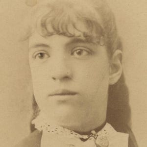 May include: A sepia-toned portrait photograph of a person with wavy hair and bangs, wearing a white lace-trimmed collar with a small circular pendant attached to a dark ribbon. The image has a soft focus and a vintage aesthetic with visible paper texture.