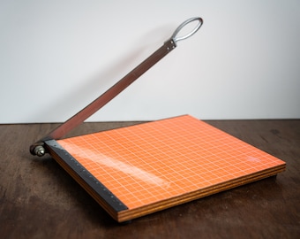 May include: A vintage orange paper cutter with a metal blade and a wooden base. The blade is raised and the cutter is sitting on a wooden surface.