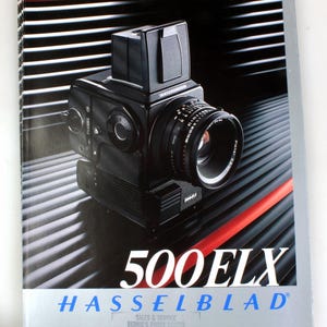 Puede incluir: Una cámara Hasselblad 500 ELX negra y plateada con una lente negra. La cámara está sobre un fondo rayado rojo y negro. El texto "500 ELX" y "HASSELBLAD" están impresos en blanco en la parte delantera de la cámara.