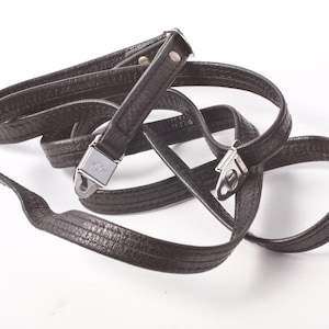 Puede incluir: Una correa de cámara de cuero negro con cierres y remaches de metal plateado. La correa tiene una textura y parece nueva. La correa está en bucle y curvada sobre un fondo blanco.