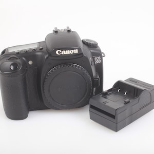 Puede incluir: Cámara réflex digital Canon EOS 20D negra con tapa de objetivo y cargador de batería negro. El logotipo de Canon está en la parte superior.