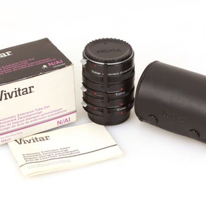 Puede incluir: Un juego de tres tubos de extensión automáticos Vivitar para cámaras Nikon, en una funda de cuero negro con una etiqueta Vivitar. Los tubos son negros con marcas rojas y la caja es blanca con texto negro.