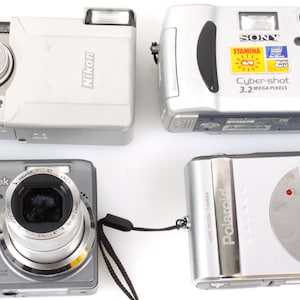 Op de afbeelding: Vier digitale camera's worden getoond tegen een witte achtergrond. De camera's zijn zilver en grijs, met merken als Nikon, Kodak, Sony Cyber-shot en Polaroid. Elke camera heeft een lens en flitser.