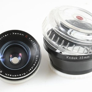 May include: A vintage camera lens with the text "Retina-Curtar-Xenon C f/4/35mm" and a black and clear Kodak 35mm lens case. The lens is black with silver accents. The case is black with a clear top.