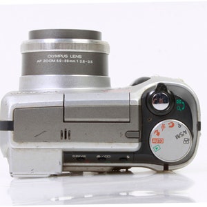 Könnte beinhalten: Eine silberne Digitalkamera mit einem schwarzen Objektiv. Das Objektiv trägt den Text "OLYMPUS LENS AF ZOOM 59-59mm 1:28-35". Die Kamera hat ein schwarzes Zifferblatt mit dem Text "AUTO, A/S/M" darauf.