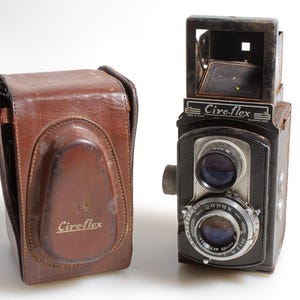 May include: A vintage Ciro-flex camera and brown leather case. The camera is black with silver detailing and the text "Ciro-flex" is visible. The brown leather case also has the "Ciro-flex" logo. The camera is a classic design.