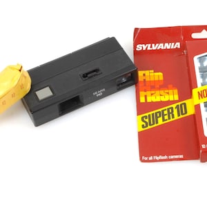 May include: A black plastic camera with a built-in flash. The camera is next to a package of Sylvania Flip Flash Super 10 flashbulbs. The package is red with yellow text and shows 10 guaranteed flashes.