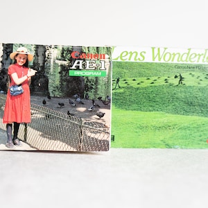 Könnte beinhalten: Zwei Bedienungsanleitungen für eine Canon AE-1 Program Kamera. Das Heft auf der linken Seite zeigt ein junges Mädchen in einem roten Kleid und einem Strohhut, das vor einem Zaun mit Tauben steht. Das Heft auf der rechten Seite zeigt ein grünes Feld mit Personen, die in der Ferne gehen, und den Text "Lens Wonderland Canon New FD Lens" in der oberen rechten Ecke.