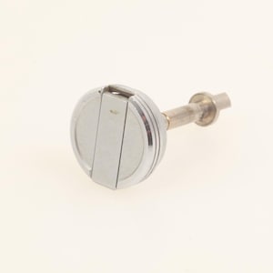 Puede incluir: Un tapón de botella plateado con una parte superior redonda y estriada y una pieza central rectangular. El tapón tiene un tallo largo y delgado y un extremo pequeño y cilíndrico. El objeto está sobre un fondo liso blanquecino.