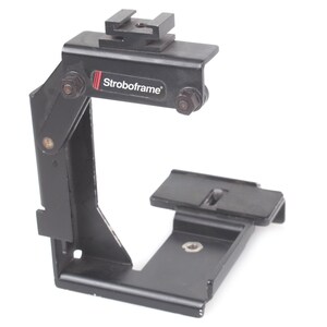 Op de afbeelding: Zwarte metalen Stroboframe camerabeugel met een bovenste schoenbevestiging en een basisplaat. De beugel heeft een verstelbare arm en de merknaam "Stroboframe" staat op de zijkant.