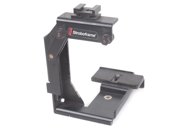 Op de afbeelding: Zwarte metalen Stroboframe camerabeugel met een bovenste schoenbevestiging en een basisplaat. De beugel heeft een verstelbare arm en de merknaam "Stroboframe" staat op de zijkant.