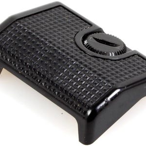 May include: A black camera accessory with a textured surface and a screw. The rectangular piece has a diamond pattern and rounded edges. The screw is circular and sits on the top of the accessory.