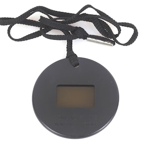 Op de afbeelding: Een cirkelvormige zwarte hanger met een rechthoekig digitaal display in het midden. De hanger is bevestigd aan een zwart geweven koord dat is voorzien van een zilverkleurige metalen punt. Het oppervlak van het zwarte ronde frame is glad en mat.