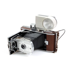 Op de afbeelding: Een vintage Polaroid Land Camera met een bruine leren hoes en een ingebouwde flitser. De camera is open en de lens is zichtbaar.
