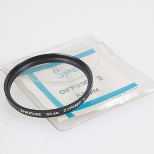 May include: A black Spiratone 55mm lens filter, with the text "Diffuser 2" and "55 mm" printed on it. The filter is in a clear plastic sleeve with a white and blue label. The label also says "Made in Japan".
