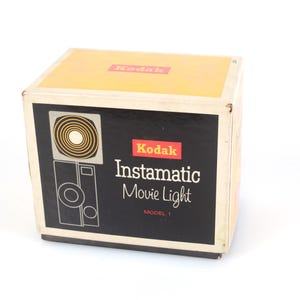 Peut inclure: Une boîte en carton rectangulaire pour un Kodak Instamatic Movie Light Model 1. La boîte présente un panneau supérieur jaune avec le logo Kodak rouge, et un panneau avant noir avec le texte blanc Kodak Instamatic Movie Light Model 1.