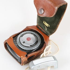 May include: A vintage black light meter with a silver circular dial featuring the GE logo, housed in a brown leather case with a green interior lining. A separate silver metal attachment with a white rectangular insert is connected by a white cord.