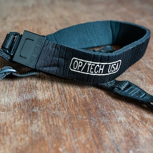 May include: Black camera strap with a padded section and a white "OP/TECH USA" logo. The strap has a black plastic buckle and a black plastic loop.