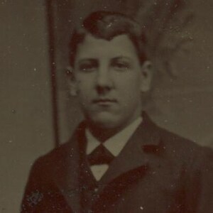 May include: Sepia-toned portrait of a person in a dark suit, white collared shirt, and a dark bow tie. The person has short, dark hair and a serious expression. The image is a vintage photograph.