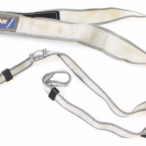 Pode incluir: Uma correia de câmera branca e cinza com uma correia ajustável preta e um logotipo Nikon azul e branco.