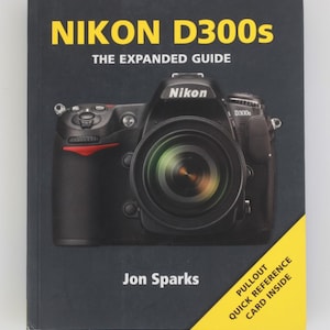 Könnte beinhalten: Ein schwarzes Buch mit dem Titel NIKON D300s THE EXPANDED GUIDE von Jon Sparks. Das Cover zeigt ein großes Foto einer schwarzen Nikon D300s Kamera mit Objektiv. Ein gelbes diagonales Eckbanner besagt PULLOUT QUICK REFERENCE CARD INSIDE.