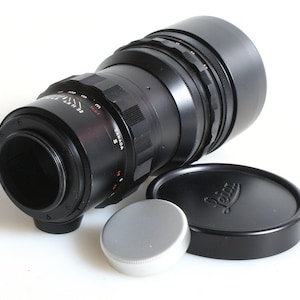 K&ouml;nnte beinhalten: Ein schwarzes Leica-Teleobjektiv mit einer silbernen Objektivkappe und einer schwarzen Objektivkappe mit dem Leica-Logo. Das Objektiv hat ein schwarzes Metallgeh&auml;use mit einem silbernen Ring um den Objektivtubus. Das Objektiv hat einen schwarzen Gummibeschlag und eine schwarze Metall-Gegenlichtblende.