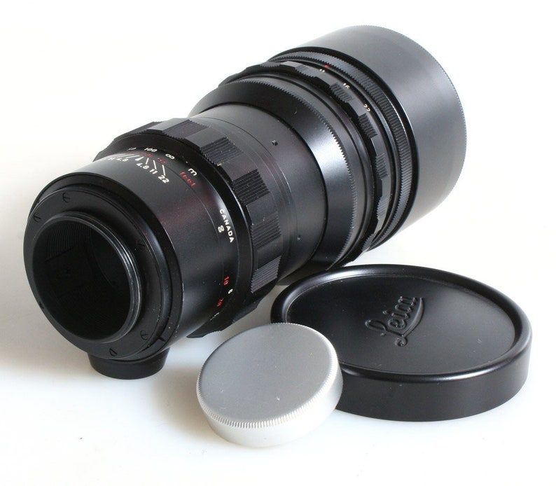 K&ouml;nnte beinhalten: Ein schwarzes Leica-Teleobjektiv mit einer silbernen Objektivkappe und einer schwarzen Objektivkappe mit dem Leica-Logo. Das Objektiv hat ein schwarzes Metallgeh&auml;use mit einem silbernen Ring um den Objektivtubus. Das Objektiv hat einen schwarzen Gummibeschlag und eine schwarze Metall-Gegenlichtblende.