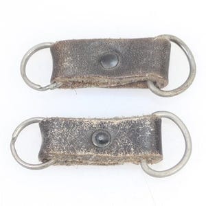 以下が含まれることがあります： 金属製のリングが付いた茶色の革製のストラップが2本。各ストラップには黒いボタンが付いています。