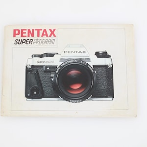 Könnte beinhalten: Eine Vintage Pentax Super Program Kamera mit einem schwarz-silbernen Gehäuse und einem großen Objektiv. Das Objektiv hat einen roten Innenraum. Die Kamera befindet sich auf einem cremefarbenen Hintergrund mit den Worten "PENTAX SUPER PROGRAM" in Rot und Schwarz.