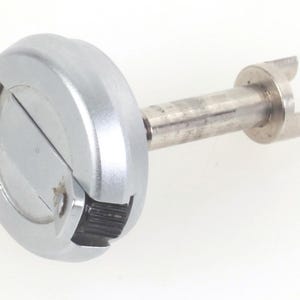 Puede incluir: Un componente mecánico plateado con una cabeza circular y un vástago cilíndrico. La cabeza tiene un diseño ranurado y el vástago termina en forma de horquilla. Probablemente parte de un mecanismo más grande.