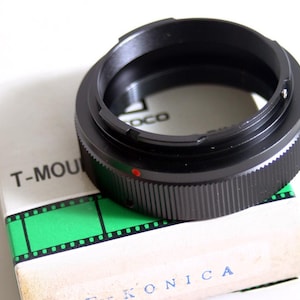 May include: Black camera lens adapter with a red dot on the side. The adapter is labeled "T-Mount" and is sitting on a box labeled "Konica".