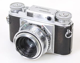 May include: A vintage Voigtlander Prominent camera with a Skopar 1:3.5/50 lens. The camera features a silver body, black leather grip, and various dials and settings. The lens is surrounded by a silver ring with measurement markings.