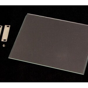 Könnte beinhalten: Eine klare, rechteckige Glasscheibe mit abgeschrägten Kanten, zusammen mit zwei silbernen Metallhalterungen und kleinen Schrauben. Die Artikel sind auf einer schwarzen Oberfläche angeordnet, was auf ein Hardware- oder Konstruktionselement hindeutet.