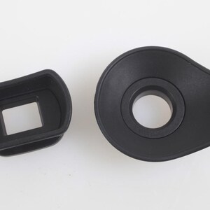 May include: Two black camera accessories, an eyepiece and a viewfinder, are displayed against a white background. The eyepiece has a rectangular opening, while the viewfinder has a circular opening. Both are made of a matte black material.