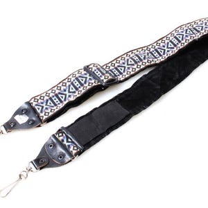 Op de afbeelding: Camerariem met een geometrisch patroon in wit, blauw en bruin. De riem heeft zilveren metalen sluitingen en een zwarte suède achterkant. Het patroon gedeelte is ongeveer 5 cm breed.
