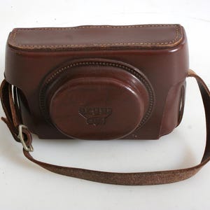 May include: A brown leather camera case with a strap. The case has a flap that covers the lens of the camera. The case has the text "Leica" embossed on the front.